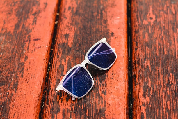 A pair of sunglasses with transparent frames rests on a weathered wooden surface. The lenses appear dark and have a reflective quality, with visible light glare. The wooden surface is painted in a reddish-brown color and shows signs of wear and aging.