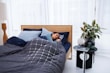 A person is sleeping comfortably in a bed with a dark gray and blue color scheme. The bedding includes a quilted comforter with a geometric pattern and multiple pillows. Next to the bed, there is a small black table holding decorative items like a plant, a vase, a book, and glasses. The environment is calm and modern, featuring sheer white curtains and a wooden floor lamp.