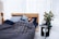 A person is sleeping comfortably in a bed with a dark gray and blue color scheme. The bedding includes a quilted comforter with a geometric pattern and multiple pillows. Next to the bed, there is a small black table holding decorative items like a plant, a vase, a book, and glasses. The environment is calm and modern, featuring sheer white curtains and a wooden floor lamp.