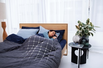 A person is sleeping comfortably in a bed with a dark gray and blue color scheme. The bedding includes a quilted comforter with a geometric pattern and multiple pillows. Next to the bed, there is a small black table holding decorative items like a plant, a vase, a book, and glasses. The environment is calm and modern, featuring sheer white curtains and a wooden floor lamp.