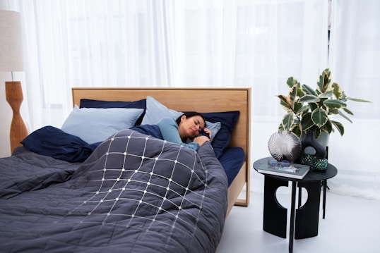 A person is sleeping comfortably in a bed with a dark gray and blue color scheme. The bedding includes a quilted comforter with a geometric pattern and multiple pillows. Next to the bed, there is a small black table holding decorative items like a plant, a vase, a book, and glasses. The environment is calm and modern, featuring sheer white curtains and a wooden floor lamp.