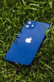 A blue smartphone, with raindrops on its surface, is lying face up on vibrant green grass. The iconic apple logo is visible in the center of the phone's back.