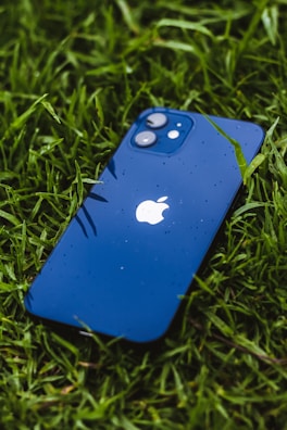 A blue smartphone, with raindrops on its surface, is lying face up on vibrant green grass. The iconic apple logo is visible in the center of the phone's back.