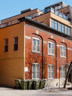A multi-story building with a mix of old and modern architecture featuring red brick and large windows on the lower floors, and tan stucco with glass railings on the upper floors. Several green recycling bins are lined up against the building. A 'No Parking' sign is visible on the pavement next to a bare tree, suggesting a winter or early spring setting. The shadows of branches are cast on the building.