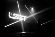 A dark concert scene with a DJ booth and a performer on stage. Spotlights and stage lights illuminate the area with beams crossing in various directions. A large logo is projected on a screen in the background.