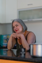 An older woman with gray hair and red lipstick is leaning on a kitchen counter, resting her chin on her hands. She is wearing a sleeveless top and an orange beaded necklace. A stainless steel pot is visible in the foreground.