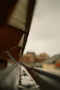 A close-up view of a rain gutter, capturing a droplet of water creating a splash as it hits the surface. The gutter is aligned with the edge of a roof, and the background appears blurred, highlighting the sharpness of the water droplet.