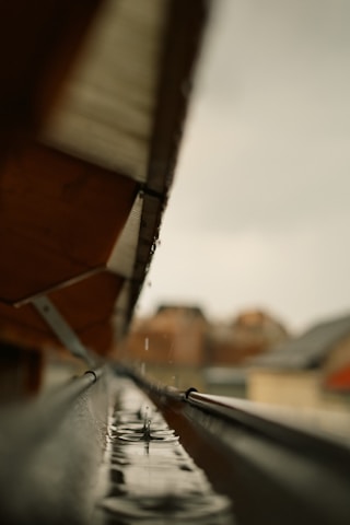 A close-up view of a rain gutter, capturing a droplet of water creating a splash as it hits the surface. The gutter is aligned with the edge of a roof, and the background appears blurred, highlighting the sharpness of the water droplet.