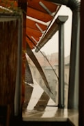 A close-up view of an outdoor area with a slanted roof, possibly made of metal and wood, with structures supporting the roof visible. Rainwater is seen on the ledge below, and a piece of mesh or netting hangs down over the side. Across the way, the blurred outlines of buildings are visible in the background.