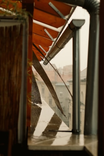 A close-up view of an outdoor area with a slanted roof, possibly made of metal and wood, with structures supporting the roof visible. Rainwater is seen on the ledge below, and a piece of mesh or netting hangs down over the side. Across the way, the blurred outlines of buildings are visible in the background.