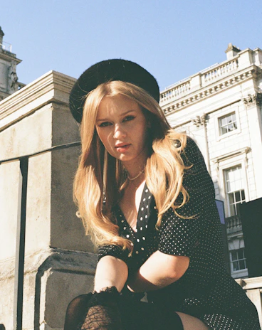 A smiling woman wearing a classic black beret, standing in a sunlit Parisian street.