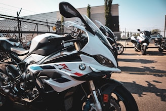 Several high-performance motorcycles are lined up in a parking lot on a sunny day. They feature sleek designs with a prominent logo on the side. The background includes a chain-link fence and some industrial buildings, contributing to an urban setting.