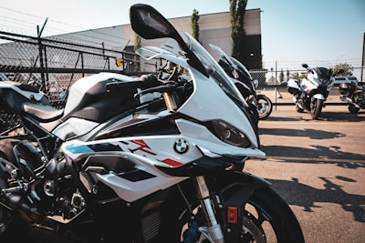Several high-performance motorcycles are lined up in a parking lot on a sunny day. They feature sleek designs with a prominent logo on the side. The background includes a chain-link fence and some industrial buildings, contributing to an urban setting.