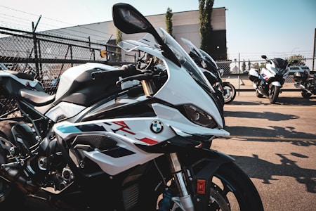 Several high-performance motorcycles are lined up in a parking lot on a sunny day. They feature sleek designs with a prominent logo on the side. The background includes a chain-link fence and some industrial buildings, contributing to an urban setting.