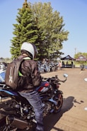 A person wearing a helmet and backpack sits on a BMW motorcycle, surrounded by other motorcycles. In the background, there are several people gathered near tents with the BMW Motorrad logo, and there is a large tree providing shade.
