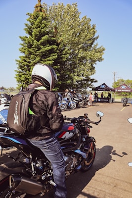 A person wearing a helmet and backpack sits on a BMW motorcycle, surrounded by other motorcycles. In the background, there are several people gathered near tents with the BMW Motorrad logo, and there is a large tree providing shade.