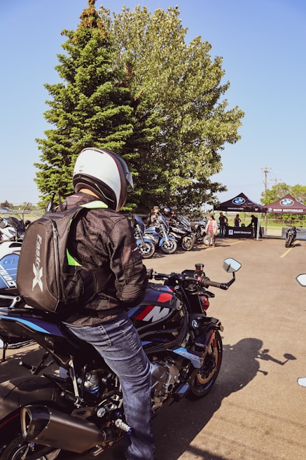 A person wearing a helmet and backpack sits on a BMW motorcycle, surrounded by other motorcycles. In the background, there are several people gathered near tents with the BMW Motorrad logo, and there is a large tree providing shade.