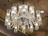 A luxurious and ornate crystal chandelier is hanging from a ceiling with a decorative design. The chandelier has multiple arms adorned with intricate glasswork and crystal pendants, each supporting a candle-like light bulb covered with an etched glass shade. The ceiling features molding and recessed lighting which adds to the opulent atmosphere.