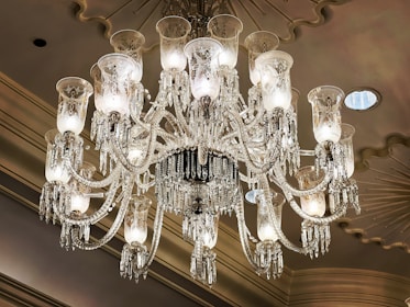 A luxurious and ornate crystal chandelier is hanging from a ceiling with a decorative design. The chandelier has multiple arms adorned with intricate glasswork and crystal pendants, each supporting a candle-like light bulb covered with an etched glass shade. The ceiling features molding and recessed lighting which adds to the opulent atmosphere.