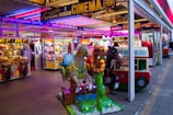 A vibrant amusement arcade featuring various games and bright neon signs. Mechanical horse rides are in the foreground, surrounded by claw machines with plush toys. The entrance is adorned with signs for a cinema and arcade, creating a lively and colorful atmosphere.