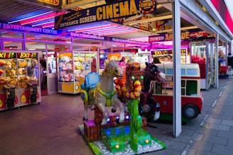A vibrant amusement arcade featuring various games and bright neon signs. Mechanical horse rides are in the foreground, surrounded by claw machines with plush toys. The entrance is adorned with signs for a cinema and arcade, creating a lively and colorful atmosphere.