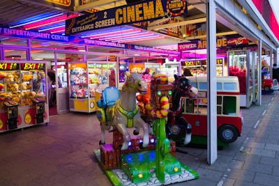 A vibrant amusement arcade featuring various games and bright neon signs. Mechanical horse rides are in the foreground, surrounded by claw machines with plush toys. The entrance is adorned with signs for a cinema and arcade, creating a lively and colorful atmosphere.