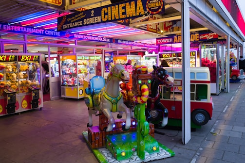 A vibrant amusement arcade featuring various games and bright neon signs. Mechanical horse rides are in the foreground, surrounded by claw machines with plush toys. The entrance is adorned with signs for a cinema and arcade, creating a lively and colorful atmosphere.