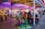 A vibrant amusement arcade featuring various games and bright neon signs. Mechanical horse rides are in the foreground, surrounded by claw machines with plush toys. The entrance is adorned with signs for a cinema and arcade, creating a lively and colorful atmosphere.