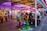 A vibrant amusement arcade featuring various games and bright neon signs. Mechanical horse rides are in the foreground, surrounded by claw machines with plush toys. The entrance is adorned with signs for a cinema and arcade, creating a lively and colorful atmosphere.