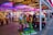 A vibrant amusement arcade featuring various games and bright neon signs. Mechanical horse rides are in the foreground, surrounded by claw machines with plush toys. The entrance is adorned with signs for a cinema and arcade, creating a lively and colorful atmosphere.