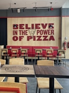 A restaurant interior featuring a large wall sign with bold red text reading 'Believe in the Power of Pizza,' incorporating a slice of pizza graphic. The seating area includes red upholstered benches and light wooden chairs with black metal frames. Pendant lights hang from the ceiling, and there are wooden tables with the restaurant's branding.