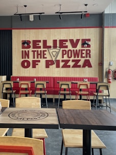 A restaurant interior featuring a large wall sign with bold red text reading 'Believe in the Power of Pizza,' incorporating a slice of pizza graphic. The seating area includes red upholstered benches and light wooden chairs with black metal frames. Pendant lights hang from the ceiling, and there are wooden tables with the restaurant's branding.