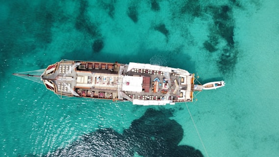 An aerial view of a large ship floating on clear turquoise water. The ship features multiple decks with seating areas and parasols, suggesting a leisurely environment. The water beneath is a pristine blue with dark patches indicating varying depths or underwater features.