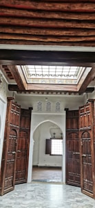 A room with intricately designed wooden doors slightly open, revealing an interior space with white walls, decorative moldings, and a geometric stained glass skylight allowing natural light to enter. The ceiling features exposed wooden beams, adding a rustic charm to the architecture.