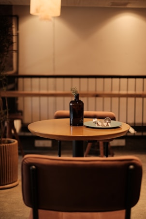 A cozy, dimly lit caf&eacute; interior with a small round table set for one. The table holds a dark glass vase with a single small flower and a plate with utensils. Warm lighting casts a gentle glow, creating a relaxed ambiance. Wooden furniture adds to the warm, inviting atmosphere.