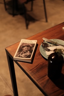 A book titled 'On Photography' by Susan Sontag is lying on a wooden table next to a set of cutlery placed on a light blue plate, and a dark-colored bottle. The photograph is taken in a dimly lit environment, giving a warm and cozy feel to the setting.