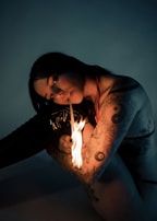 A tattooed woman is holding a lit matchstick close to her face. The dim, moody lighting accentuates her features and the detailed tattoos on her arm. She has dark hair and is wearing minimal clothing, suggesting an intimate, contemplative moment.