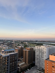 A cityscape featuring a blend of modern high-rise buildings and expansive urban planning. The horizon showcases a distant skyline under a blue sky with soft pastel hues from a setting or rising sun. Major roads and a waterway are visible, adding to the urban vibe.