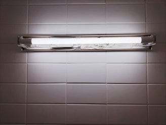 A fluorescent light fixture is mounted on a white tiled wall. The light is turned on, casting a bright, even glow across the surrounding tiles. The fixture contains two fluorescent tubes, enclosed in a metallic frame.