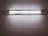 A fluorescent light fixture is mounted on a white tiled wall. The light is turned on, casting a bright, even glow across the surrounding tiles. The fixture contains two fluorescent tubes, enclosed in a metallic frame.