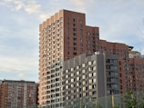 A cluster of modern, high-rise apartment buildings with a mix of beige and gray facades. The structures are rectangular with multiple balconies and large windows. The sky is partially cloudy, adding a soft, pastel background.
