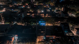 A nighttime aerial view of an urban neighborhood with buildings densely packed together, illuminated by streetlights and lights from windows. The scene is enveloped in darkness, with scattered bright spots that highlight various structures.