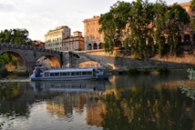 A river cruise boat sails along a calm waterway, reflecting the late afternoon sunlight. To the left, a stone bridge spans the river, with its large arches mirrored on the water's surface. On the banks, tall, lush trees provide shade and frame the scene with vibrant greenery. Historic buildings with warm-colored facades stand in the background, casting a golden glow.