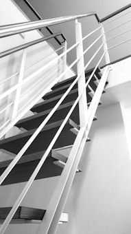 A black and white photograph featuring a modern indoor staircase with sleek, minimalist design. The staircase is characterized by thin metal railings and evenly spaced steps, creating a geometric and contemporary aesthetic. The perspective is angled upward, emphasizing the clean lines and architectural elements.