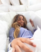 A person lying comfortably surrounded by fluffy white pillows, wearing a blue and white striped shirt, and smiling warmly.