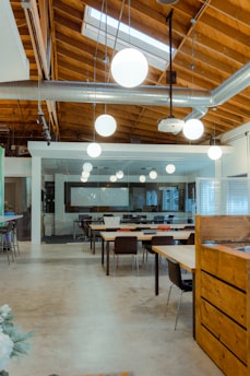 An open office space with modern design elements including exposed wooden beams and ductwork. There are several tables and chairs arranged for collaborative work, with pendant lights hanging from the ceiling. In the background, a glass wall separates the space from another area.
