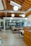 An open office space with modern design elements including exposed wooden beams and ductwork. There are several tables and chairs arranged for collaborative work, with pendant lights hanging from the ceiling. In the background, a glass wall separates the space from another area.