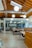 An open office space with modern design elements including exposed wooden beams and ductwork. There are several tables and chairs arranged for collaborative work, with pendant lights hanging from the ceiling. In the background, a glass wall separates the space from another area.