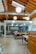 An open office space with modern design elements including exposed wooden beams and ductwork. There are several tables and chairs arranged for collaborative work, with pendant lights hanging from the ceiling. In the background, a glass wall separates the space from another area.
