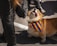 A dog wearing a colorful rainbow bandana appears engaged and animated, seeming to bark or howl. The scene includes people wearing black clothing, including boots and a faux fur sleeve, suggesting participation in an outdoor event.
